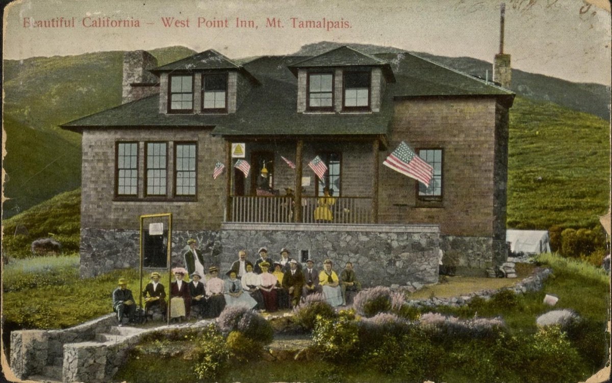 Hand-coloured postcard of the West Point Inn decorated with American flags, with visitors seated in front