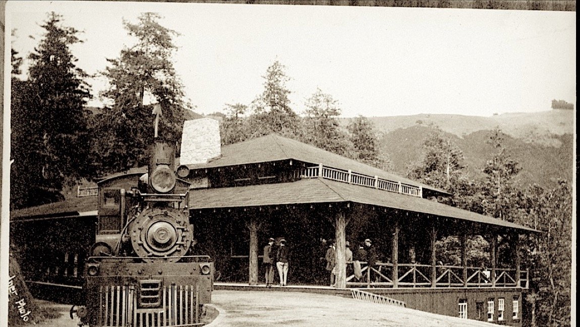 A steam engine in front of a long open-sided lodge among redwoods