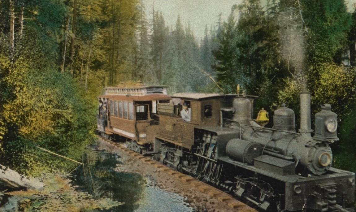 A hand-coloured postcard of a geared steam engine pushing a passenger car beside a creek in the redwoods