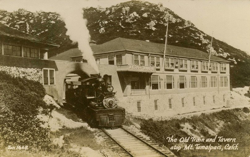 Postcard: a steam engine emerging from the arched bridge beside the Tavern of Tamalpais, with the summit rocks behind